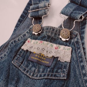 May include: Blue denim overalls featuring a chest pocket with a white lace trim adorned with a small pink floral pattern. The garment includes metal hardware, adjustable shoulder straps, and a blue fabric label reading OshKosh B'Gosh The Genuine Article.