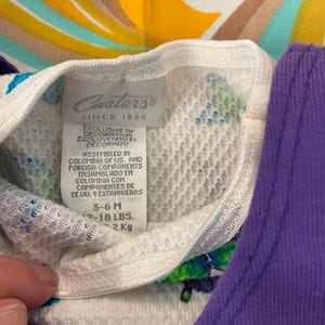 May include: White Carter's brand baby onesie with a colorful swirl pattern. The tag says "Exclusive of decoration, Excluyendo el decorado." and "Assembled in Colombia of US. and foreign components, Ensamblado en Colombia con componentes de EE.UU. y extranjeros." The tag also says "3-6M, 12-18 LBS, 2 KG."