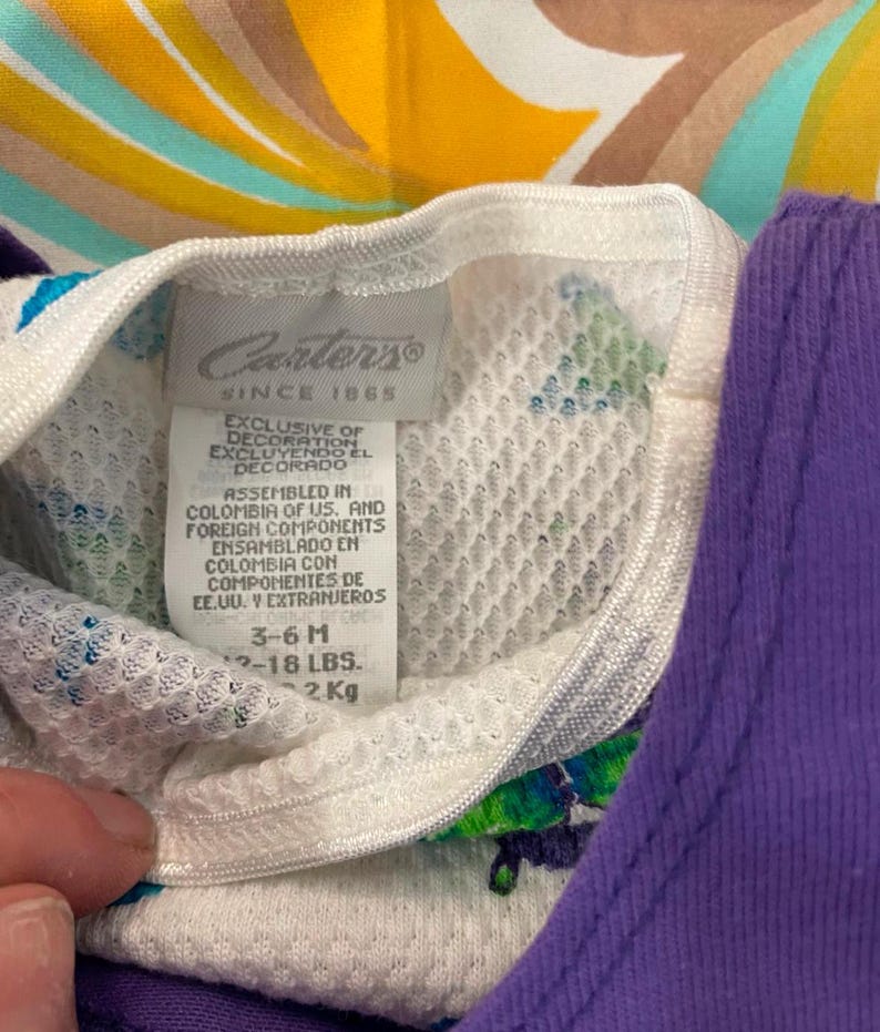 May include: White Carter's brand baby onesie with a colorful swirl pattern. The tag says "Exclusive of decoration, Excluyendo el decorado." and "Assembled in Colombia of US. and foreign components, Ensamblado en Colombia con componentes de EE.UU. y extranjeros." The tag also says "3-6M, 12-18 LBS, 2 KG."