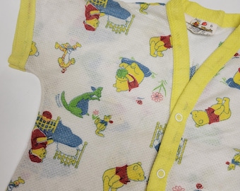 May include: A white baby onesie with a yellow trim and a Winnie the Pooh print. The onesie has a button closure and is made of a soft, comfortable fabric. The print features Winnie the Pooh, Tigger, Piglet, Eeyore, and Kanga.