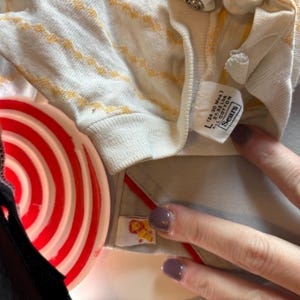 K&ouml;nnte beinhalten: Nahaufnahme eines Vintage-Babyhemdes mit cremefarbenem Grund und gelbem Zickzackmuster. Das Hemd hat ein Etikett mit der Aufschrift "L (24-30 Mos.) 27.32 Lbs. ALL COTTON Sears". Ein rot-wei&szlig; gestreifter Gegenstand ist im Vordergrund.