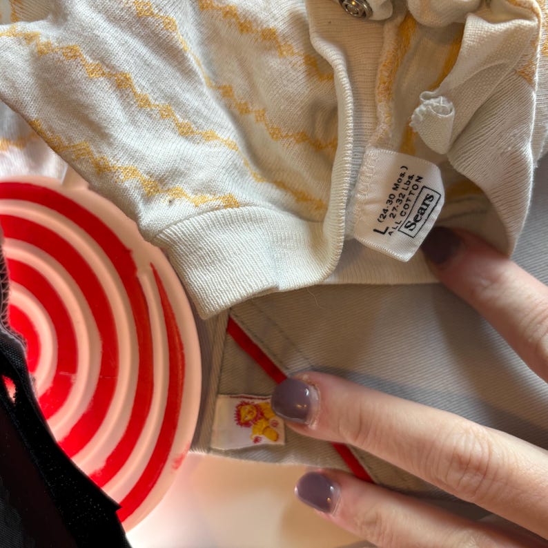 K&ouml;nnte beinhalten: Nahaufnahme eines Vintage-Babyhemdes mit cremefarbenem Grund und gelbem Zickzackmuster. Das Hemd hat ein Etikett mit der Aufschrift "L (24-30 Mos.) 27.32 Lbs. ALL COTTON Sears". Ein rot-wei&szlig; gestreifter Gegenstand ist im Vordergrund.