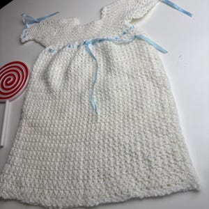K&ouml;nnte beinhalten: Wei&szlig;es geh&auml;keltes Babykleid mit angen&auml;hten Fl&uuml;geln und hellblauen B&auml;ndern. Das Kleid ist handgefertigt mit einem strukturierten Stich und hat einen runden Ausschnitt. Ein rot-wei&szlig;es Lutscher befindet sich im Hintergrund.