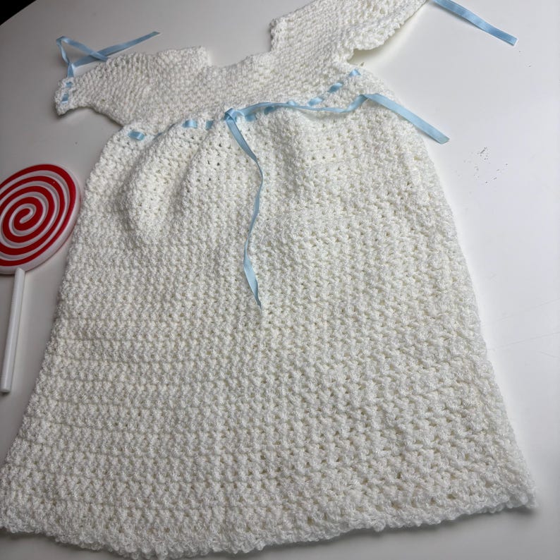 K&ouml;nnte beinhalten: Wei&szlig;es geh&auml;keltes Babykleid mit angen&auml;hten Fl&uuml;geln und hellblauen B&auml;ndern. Das Kleid ist handgefertigt mit einem strukturierten Stich und hat einen runden Ausschnitt. Ein rot-wei&szlig;es Lutscher befindet sich im Hintergrund.