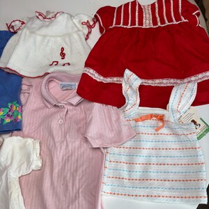 K&ouml;nnte beinhalten: Eine Auswahl an Kinderkleidung, darunter ein wei&szlig;es Kleid mit roter Verzierung und Notenstickerei, ein rotes Kleid mit wei&szlig;er Spitze, ein rosa Hemd mit Kragen und ein gestreiftes Oberteil mit einer orangefarbenen Schleife.