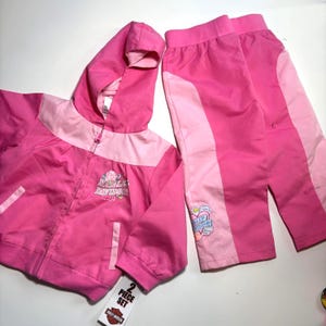 May include: A two-piece pink Harley Davidson outfit for children. The set includes a hooded jacket with a zipper and matching pants. The jacket and pants have light pink accents and Harley Davidson logos. The set is new with tags.