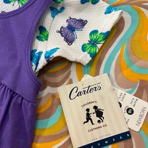 May include: Purple sleeveless dress with a white butterfly print top. The tag reads "Carter's Children's Clothing Co. 100% Cotton Playwear" and includes size information for 9 months, 9 cm, 12-18 lbs, 5.5-8 kg, 62-68 cm, 18-21 lbs, 8-9.5 kg.