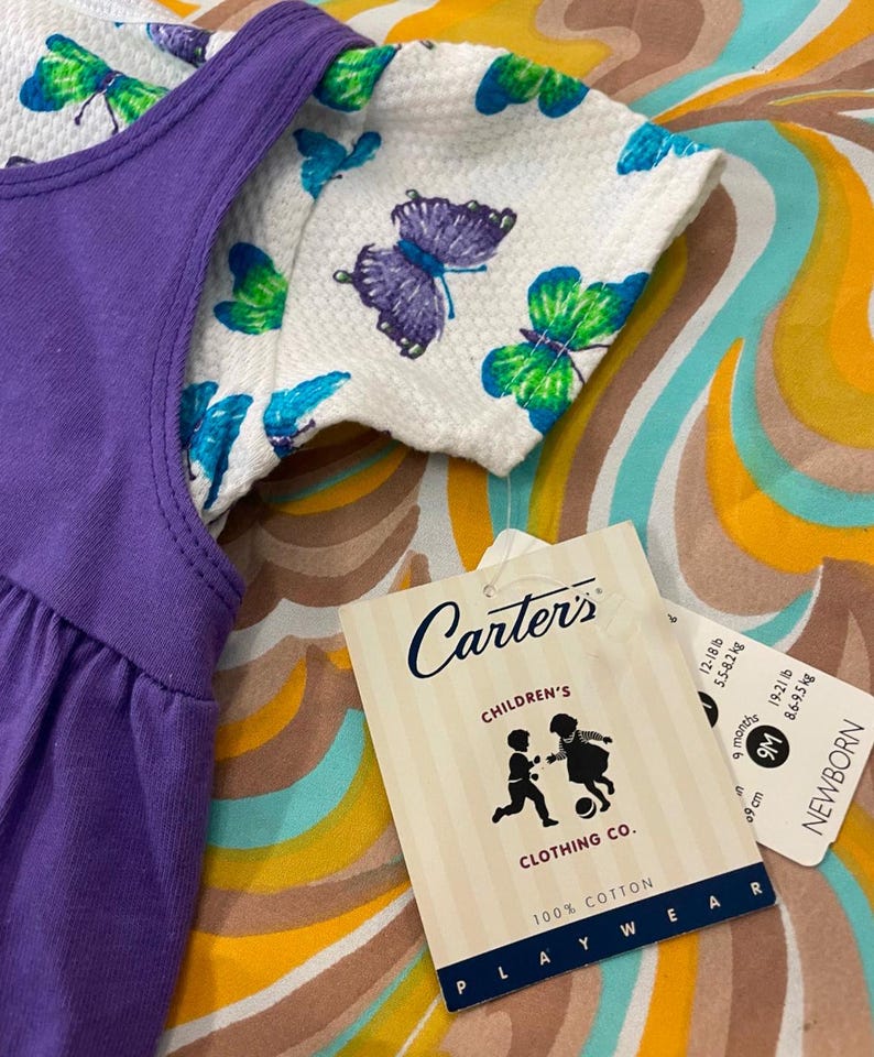 May include: Purple sleeveless dress with a white butterfly print top. The tag reads "Carter's Children's Clothing Co. 100% Cotton Playwear" and includes size information for 9 months, 9 cm, 12-18 lbs, 5.5-8 kg, 62-68 cm, 18-21 lbs, 8-9.5 kg.