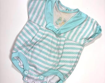 May include: A light blue and white striped baby onesie with a bow and a small embroidered design on the front. The onesie has short sleeves and snaps at the bottom.