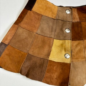 Op de afbeelding: Een bruine suède minirok met een patchwork ontwerp. De rok heeft een knoopsluiting en is gemaakt van verschillende tinten bruin suède.