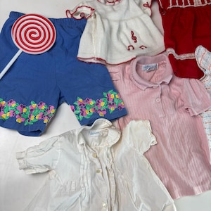K&ouml;nnte beinhalten: Eine Sammlung von Kinderkleidung, darunter blaue Shorts mit Blumenstickerei, ein wei&szlig;es Oberteil mit Notenstickerei, ein rosa Hemd mit Kragen und ein wei&szlig;es Hemd mit Kn&ouml;pfen. Ein rot-wei&szlig;es Lollipop ist ebenfalls vorhanden.