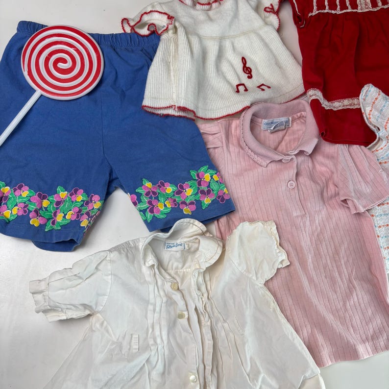 K&ouml;nnte beinhalten: Eine Sammlung von Kinderkleidung, darunter blaue Shorts mit Blumenstickerei, ein wei&szlig;es Oberteil mit Notenstickerei, ein rosa Hemd mit Kragen und ein wei&szlig;es Hemd mit Kn&ouml;pfen. Ein rot-wei&szlig;es Lollipop ist ebenfalls vorhanden.