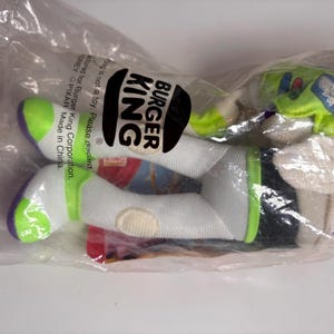 May include: A green and white plush toy of Buzz Lightyear from the Toy Story franchise. The toy is packaged in a clear plastic bag with a Burger King logo. The bag also includes the text "Bag is not a toy. Please discard.  Manufactured for Burger King Corporation.  DISNEY PIXAR Made in China."