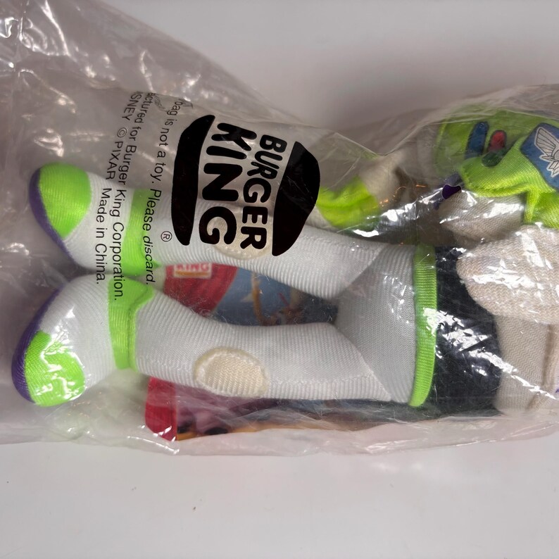 May include: A green and white plush toy of Buzz Lightyear from the Toy Story franchise. The toy is packaged in a clear plastic bag with a Burger King logo. The bag also includes the text "Bag is not a toy. Please discard.  Manufactured for Burger King Corporation.  DISNEY PIXAR Made in China."