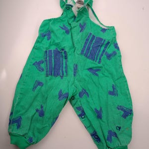 May include: A bright green overall featuring a repeating dark blue brushstroke pattern. The garment includes two front pockets with vertical blue stripes, shoulder straps with snap closures, and additional snap closures along the inner leg seams.