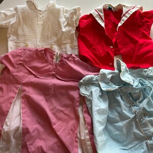 K&ouml;nnte beinhalten: Eine Sammlung von Vintage-Kinderkleidung, darunter eine wei&szlig;e Bluse mit Falten und Kn&ouml;pfen, ein rotes Oberteil mit wei&szlig;em Kragen, ein rosa Kleid mit Rei&szlig;verschluss und eine hellblaue Bluse mit Spitzenbesatz. Die Kleidungsst&uuml;cke sind auf einer wei&szlig;en Oberfl&auml;che angeordnet.