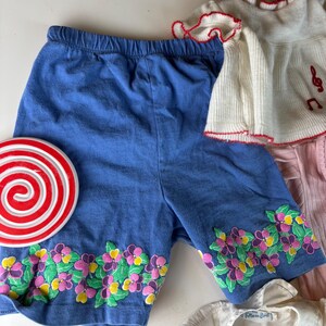 K&ouml;nnte beinhalten: Blaue Shorts mit elastischem Bund und floraler Stickerei am Saum. Die Stickerei zeigt rosa, gelbe und violette Blumen mit gr&uuml;nen Bl&auml;ttern. Ein gro&szlig;er rot-wei&szlig;er Lutscher befindet sich auf der linken Seite.