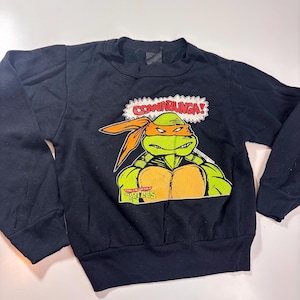 Może przedstawiać: Czarna bluza z nadrukiem Teenage Mutant Ninja Turtles. Nadruk przedstawia zieloną ninja-żółwia z pomarańczową bandaną, czerwoną maską i słowami "Cowabunga!" w dymku. Tekst "Teenage Mutant Ninja Turtles" jest również widoczny na nadruku.