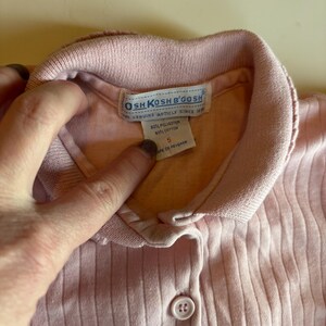 K&ouml;nnte beinhalten: Nahaufnahme eines hellrosa gerippten Strickhemdes mit Kragen. Das Hemd hat einen Knopfverschluss und ein Etikett mit der Aufschrift "OSHKOSH B'GOSH THE GENUINE ARTICLE SINCE 1895". Das Etikett gibt an, dass das Hemd aus 50 % Polyester und 50 % Baumwolle besteht.