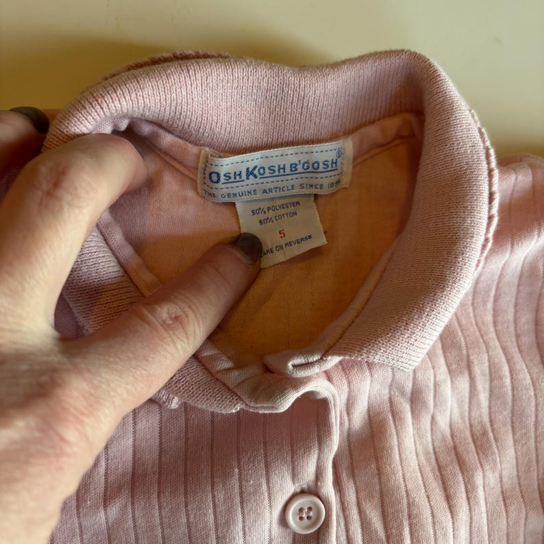 K&ouml;nnte beinhalten: Nahaufnahme eines hellrosa gerippten Strickhemdes mit Kragen. Das Hemd hat einen Knopfverschluss und ein Etikett mit der Aufschrift "OSHKOSH B'GOSH THE GENUINE ARTICLE SINCE 1895". Das Etikett gibt an, dass das Hemd aus 50 % Polyester und 50 % Baumwolle besteht.