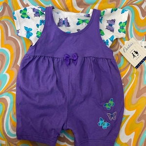 May include: A purple overall romper with butterfly embroidery and a white short-sleeved shirt with butterfly print. The outfit is labeled "Carter's" and "Onesies&reg;".