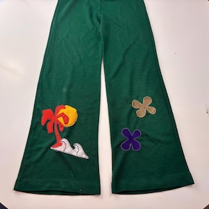 May include: Green flared trousers with colourful appliqués. The trousers feature a palm tree and sun design, along with two flower-shaped patches. The fabric appears to be a soft knit material.