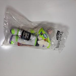 May include: A green and white plush toy of Buzz Lightyear from the movie Toy Story. The toy is packaged in a clear plastic bag with a Burger King logo and the words "Toy Story" printed on it.
