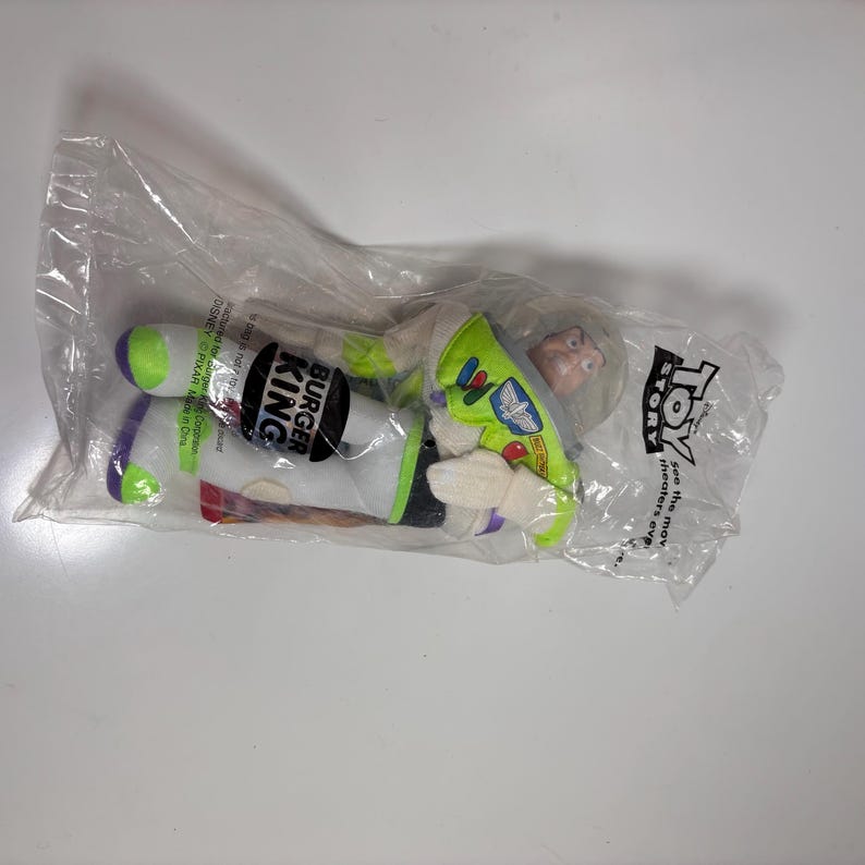 May include: A green and white plush toy of Buzz Lightyear from the movie Toy Story. The toy is packaged in a clear plastic bag with a Burger King logo and the words "Toy Story" printed on it.