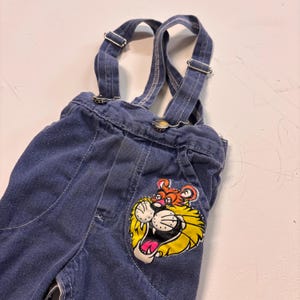 May include: Blue denim overalls with adjustable straps and metal hardware. A colorful lion graphic is on the front pocket. The overalls have a classic design, suitable for children.