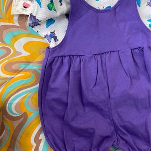 May include: A purple overall dress with a white short-sleeved shirt featuring a butterfly print. The shirt has a white background with colorful butterflies.