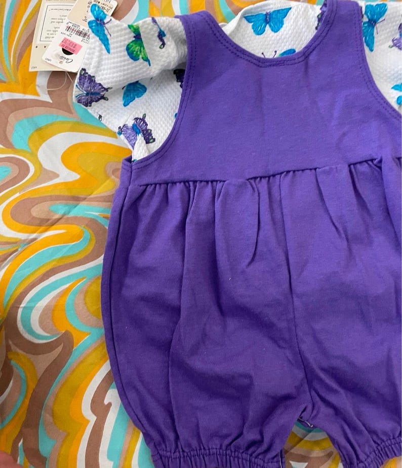 May include: A purple overall dress with a white short-sleeved shirt featuring a butterfly print. The shirt has a white background with colorful butterflies.