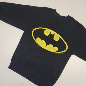 Può includere: Felpa nera con un logo di Batman giallo. Il logo è una silhouette di pipistrello stilizzata all'interno di un ovale. Il testo "TM & DC Comics Inc. 1992" è stampato sotto il logo.