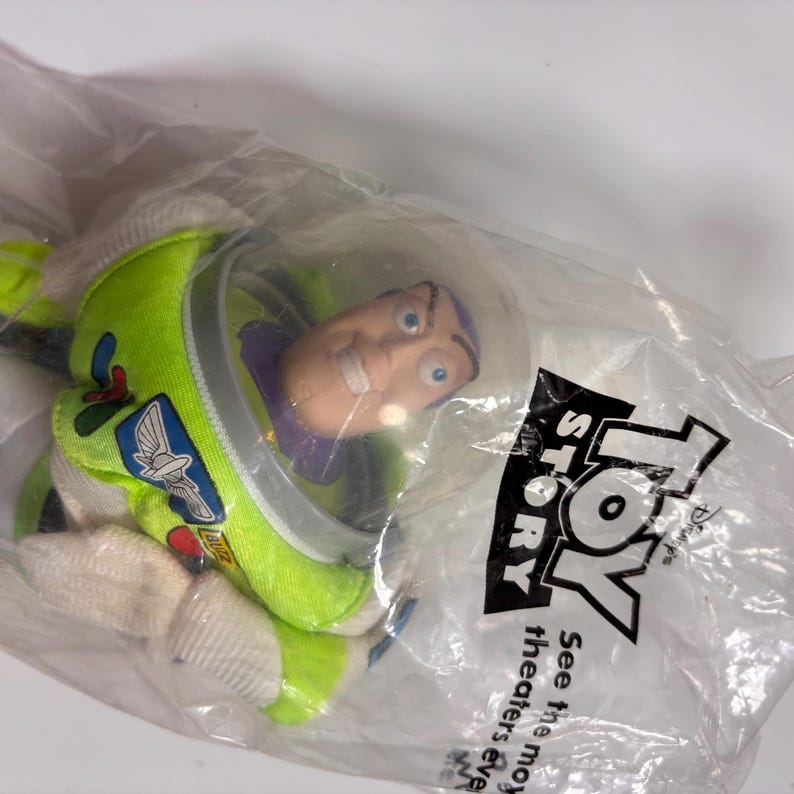 May include: A green and white plush toy of Buzz Lightyear from the movie Toy Story. The toy is in a plastic bag with the Toy Story logo and the text "See the movie in theaters everywhere".