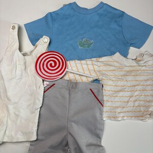 K&ouml;nnte beinhalten: Eine Auswahl an Kinderkleidung und ein Lolli. Die Kleidung umfasst eine wei&szlig;e Cord-Latzhose, ein hellblaues T-Shirt mit gr&uuml;nem Applikation, graue Shorts mit roter Paspelierung und ein gestreiftes Langarmshirt.