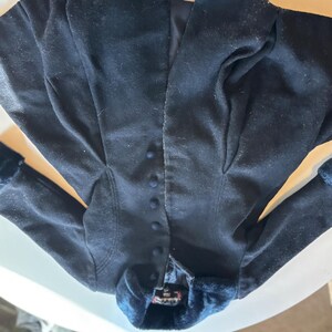 K&ouml;nnte beinhalten: Dunkelmarineblaue Jacke mit tailliertem Schnitt. Die Jacke hat eine Reihe kleiner, passender Kn&ouml;pfe auf der Vorderseite und eine pl&uuml;schige, dunkelblaue Verzierung um Kragen und Manschetten. Der Stoff scheint weich zu sein, m&ouml;glicherweise Samt.