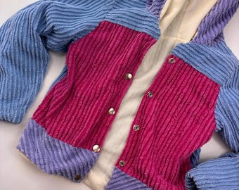May include: A colorful corduroy jacket with a hood. The jacket features a patchwork design with panels of blue, pink, and purple. It has a button-up front and a soft, cream-colored lining. The jacket is a unique and stylish piece of outerwear.