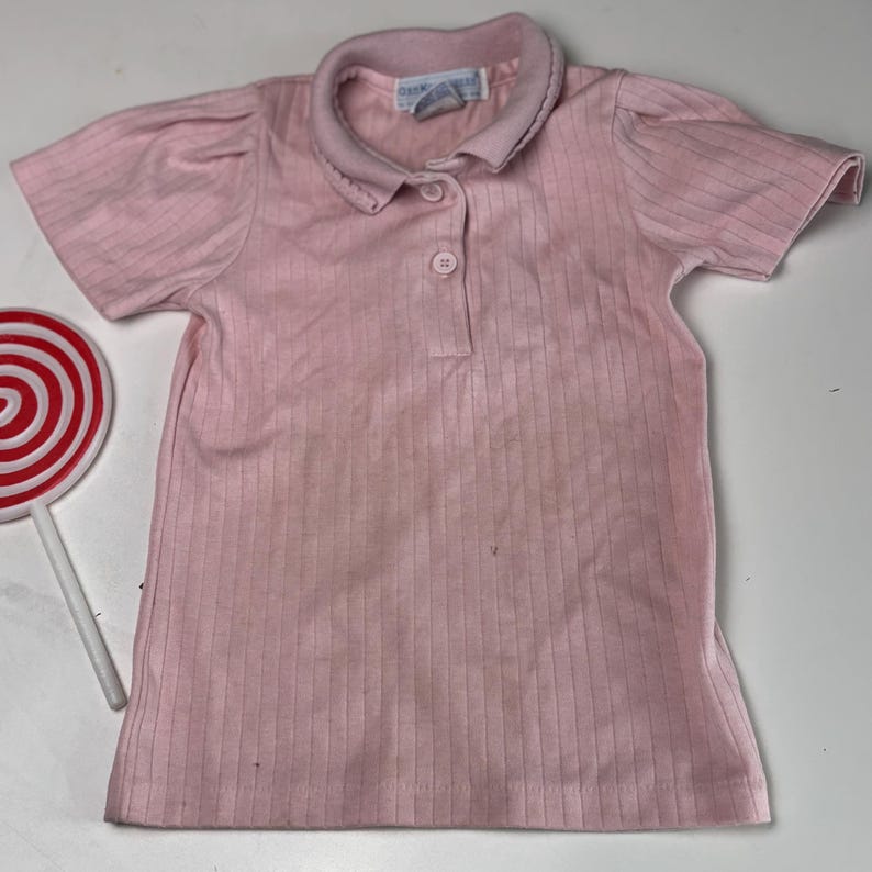 K&ouml;nnte beinhalten: Ein hellrosa geripptes Poloshirt mit kurzen &Auml;rmeln und einem Kragen. Das Shirt hat eine Knopfleiste mit drei Kn&ouml;pfen und ein kleines Etikett am Kragen. Ein rot-wei&szlig;es Lollipop ist auf der linken Seite sichtbar.