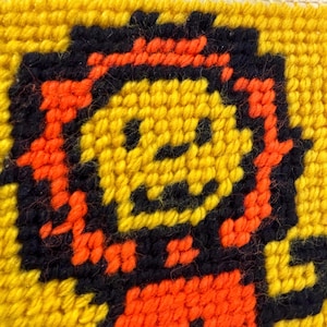 May include: A close-up of a cross-stitch design featuring a smiling character. The character is primarily yellow with black and orange accents, set against a yellow background. The design is made with yarn.