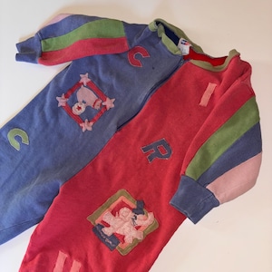May include: A vintage children's jumpsuit in blue and red. The two-piece garment has a zip down the front and colourful patches with letters and cartoon characters. The sleeves have striped accents in pink, green, and blue.