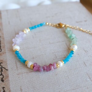 May include: A delicate bracelet with a gradient of pastel pink, blue, and green gemstone beads. The bracelet is accented with small gold beads and white pearl beads.