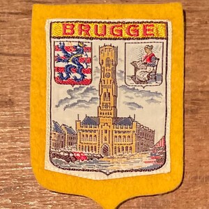 Op de afbeelding: Een gele vilten souvenirpatch in schildvorm. De patch toont het woord "BRUGGE" bovenaan, met illustraties van een toren, wapenschild en een vrouw. De achtergrond is een gedetailleerde weergave van een stadsgezicht.