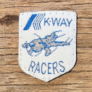 Op de afbeelding: Een wit stoffen embleem met een blauw en grijs vliegtuigontwerp en de tekst "KWAY RACERS". Het embleem is schildvormig en ligt op een houten oppervlak.