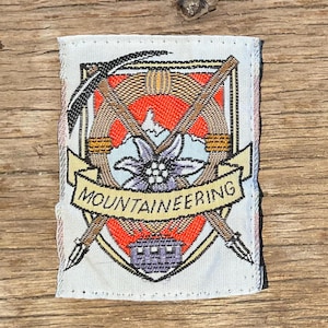 Peut inclure: Écusson rectangulaire avec un emblème d'alpinisme. Le motif comprend des piolets croisés, une fleur et le mot "MOUNTAINEERING" sur une bannière. L'écusson a un fond blanc avec des accents rouges, orange et violets.