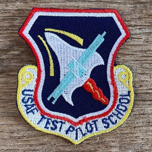 Op de afbeelding: Geborduurde patch met de tekst "USAF TEST PILOT SCHOOL" in wit en geel. De patch heeft een marineblauwe achtergrond met een wit en lichtblauw ontwerp. Het is omrand met rode, witte en gele stiksels, waarschijnlijk een militair insigne.