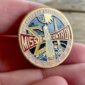 Op de afbeelding: Een ronde, goudkleurige metalen pin met de woorden "MISSION CONTROL" in rood en wit. De pin heeft een raketontwerp en de tekst "RES GESTA PAR EXCELLENTIAM" in een cirkelvormige rand. De pin wordt in een hand gehouden.