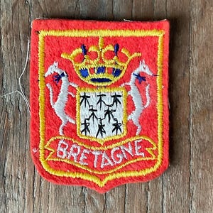 May include: A red embroidered patch with a yellow border. The patch features a crest with a crown, two white unicorns, and the word "BRETAGNE" in yellow. The crest is on a wooden surface.