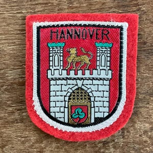 Op de afbeelding: Een rode patch met een witte rand met een wapen met het woord "HANNOVER" bovenaan. Het wapen toont een gouden leeuw op een kasteel met een groen en goud embleem aan de onderkant.