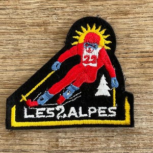 Può includere: Toppa ricamata raffigurante uno sciatore con tuta da sci rossa, con il numero 23, che scia giù per una pista. La toppa ha uno sfondo nero con la scritta "LES 2 ALPES" in bianco e dettagli gialli. Un disegno a forma di sole è sopra lo sciatore.