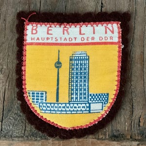 May include: A shield-shaped fabric patch with a dark brown scalloped border. The center features a yellow background with a blue graphic of the Berlin TV tower and buildings. The top section is white with the red text BERLIN HAUPTSTADT DER DDR.