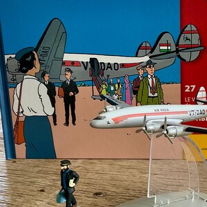 Op de afbeelding: Een modelvliegtuig met de tekst "AIR INDIA VT-DAD" wordt getoond voor een boek met een blauwe kaft en de tekst "EN AVION TINTIN". Er is ook een klein beeldje van een man in een zwart pak aanwezig.